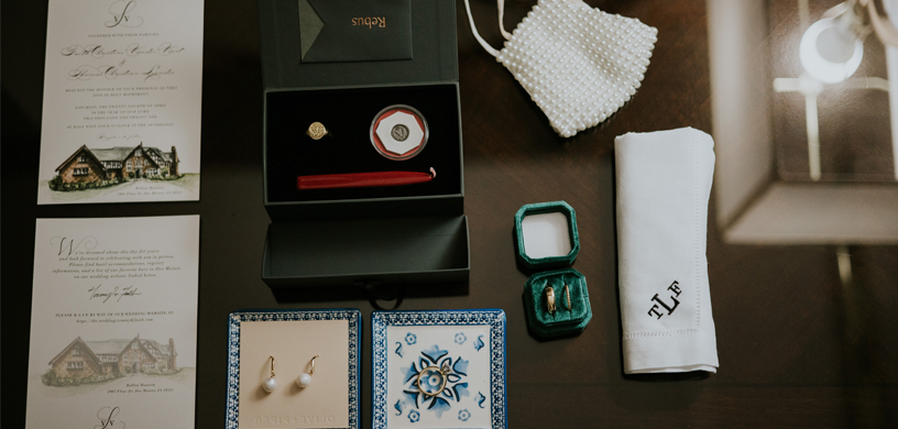 Aerial photograph of wedding paraphernalia including pearl earrings, gold signet ring, embroidered handkerchief, and wedding invitation 
